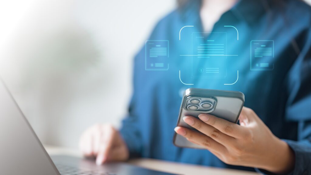 Person holding a smartphone while working on a laptop, with a digital interface suggesting document or expense scanning.