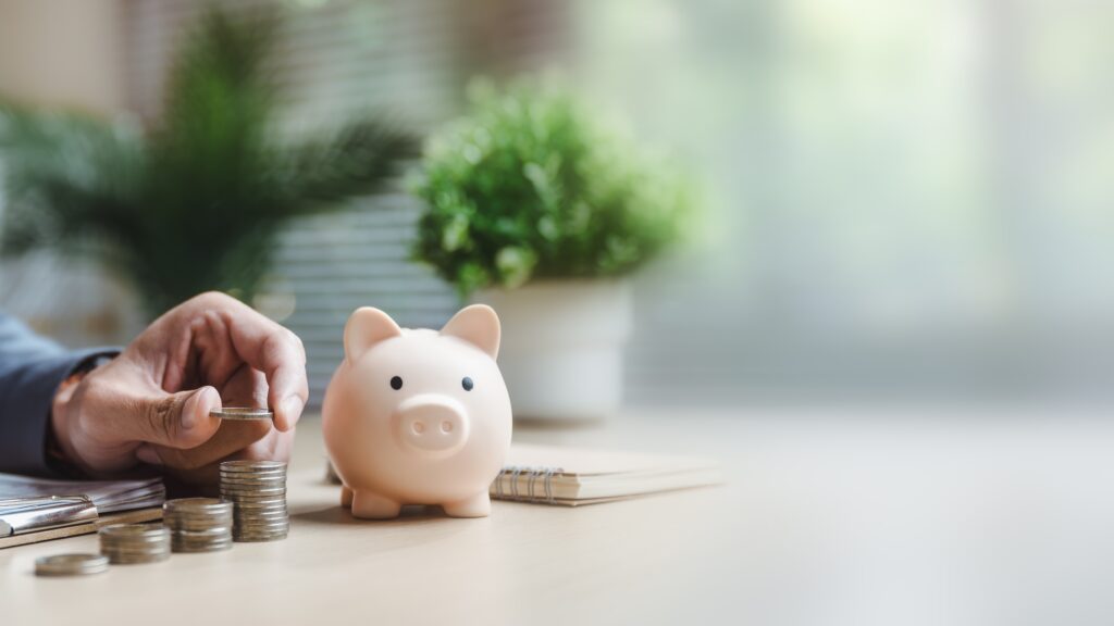 Hand placing coins next to a piggy bank on a desk, symbolising business savings and financial growth